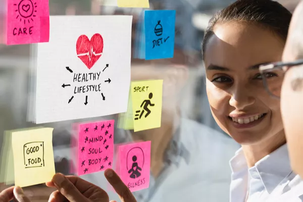 woman and second person planning with sticky notes that read, "care, good food, healthy lifestyle, mind soul body, diet, sport, wellness" 